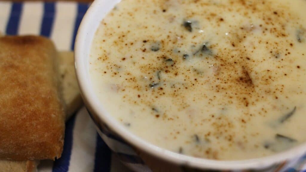 A bowl of creamy sausage potato kale soup with herbs sprinkled on top sits next to a piece of bread on a blue and white striped napkin.