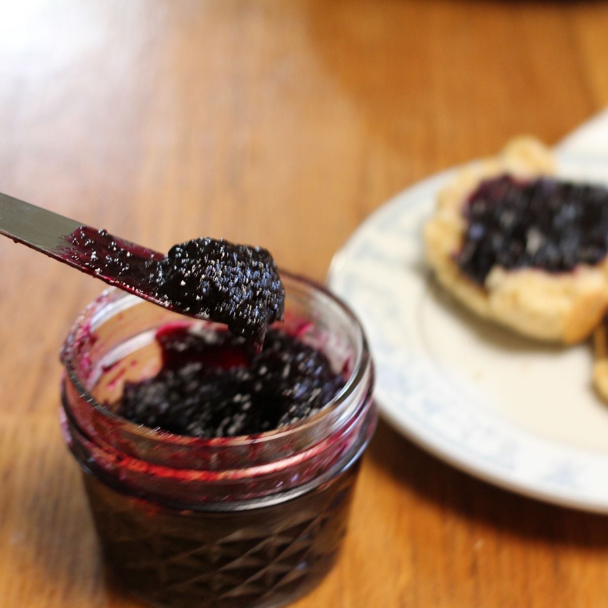A knife spreading rich grape jam from a small glass jar onto toast on a plate, set on a wooden table.