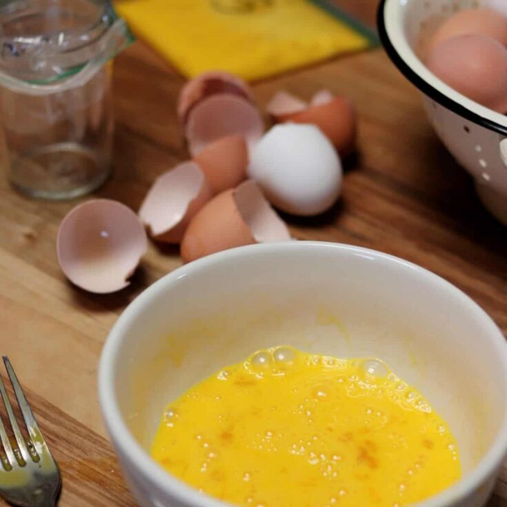 whisked eggs in a white bowl on a wooden table surrounded by egg shells, colander with farm fresh eggs, fork, mason jar lined with ziplock bag and a ziplock bag with whisked eggs for freezing