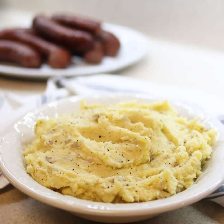 A bowl of creamy mashed potatoes seasoned with pepper. In the background, a plate with what appears to be cooked sausages is slightly out of focus. The setting is a light-colored countertop with a cloth napkin nearby.
