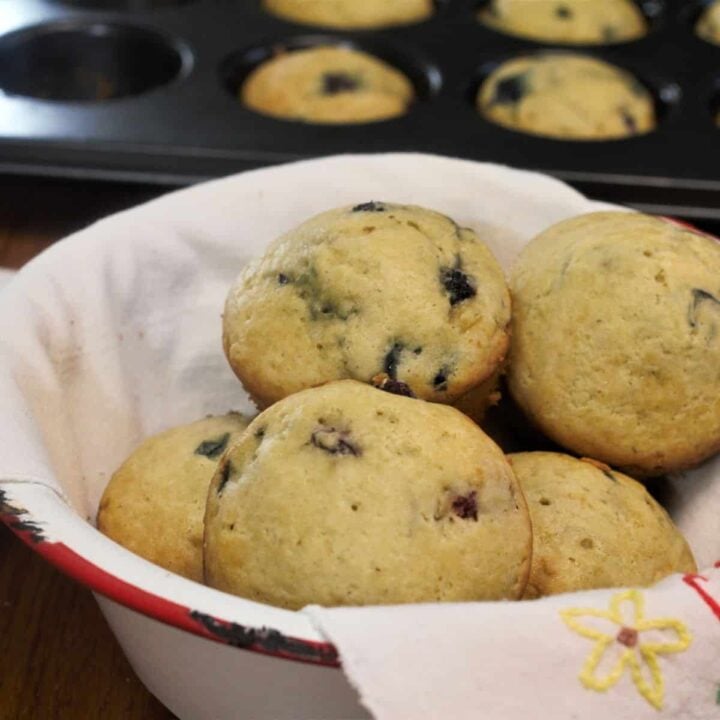 A bowl filled with freshly baked muffins, featuring visible blueberries, sits on a table. In the background, a muffin tray with more muffins is partially visible. The bowl is lined with a cloth decorated with a simple yellow star design.