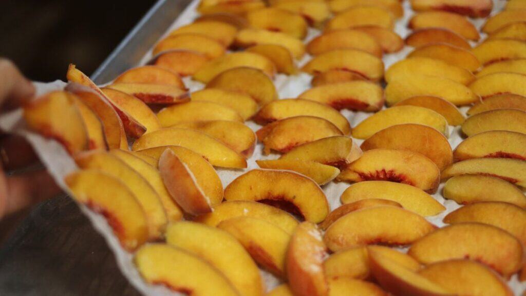 Slices of fresh peaches are arranged in neat rows on a baking sheet lined with parchment paper, ready for drying or baking. A hand is lifting the edge of the parchment paper.