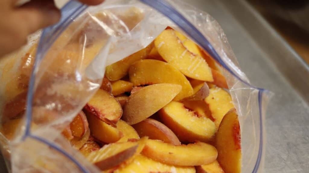 A clear plastic freezer bag filled with sliced peaches, some with reddish edges, sits on a metal surface.