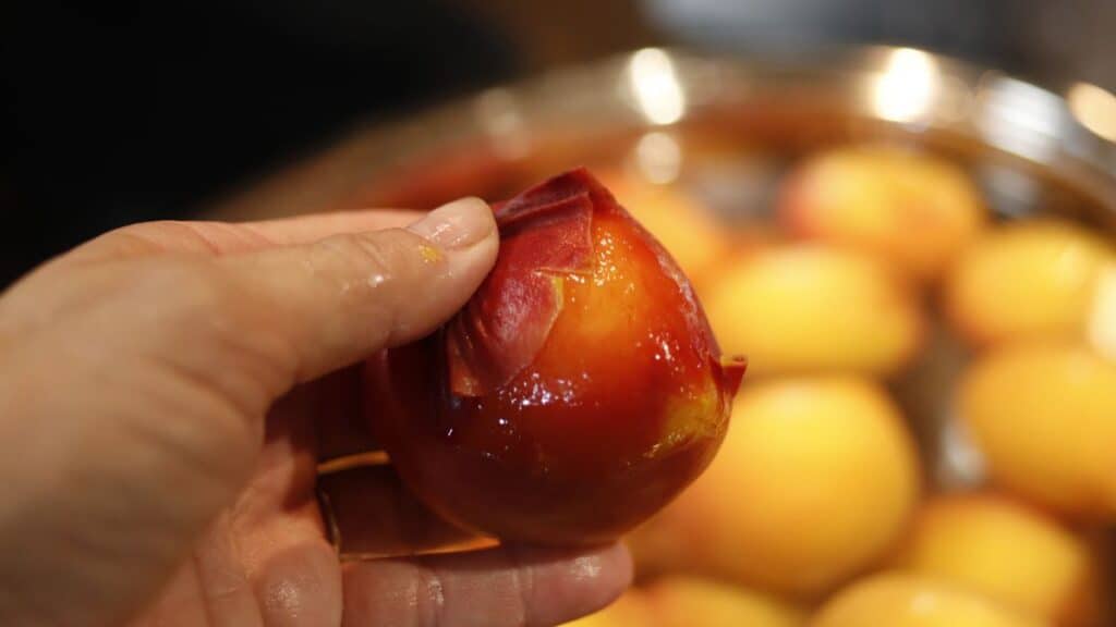 A hand is peeling the skin off a ripe nectarine. In the background, there are more nectarines in a metal bowl.