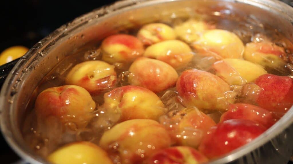 Several whole peaches are submerged in a large pot of boiling water, with bubbles forming around the fruit as they are being blanched.