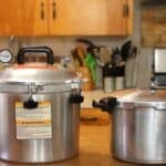 Two large metal pressure canners sit on a wooden kitchen counter; one has a dial gauge and warning labels, the other has a simple handle and knob. Kitchen utensils and cabinets are visible in the background.