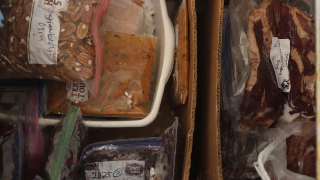 Several labeled bags of frozen food items, including baked goods, meat, and other dishes, are organized in a packed freezer drawer, with handwritten labels visible on some bags.