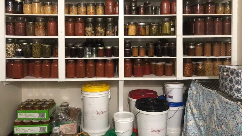 Shelves filled with assorted jars of preserved foods, with labeled buckets for popcorn, sugar, and other bulk items below. Boxes, jars, and a patterned cloth are also visible near the buckets.