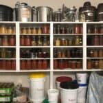 Shelves filled with jars of preserved food, such as pickles and sauces, are organized above large labeled buckets of popcorn and sugar in a pantry. Various kitchen supplies and appliances are on the top shelves.