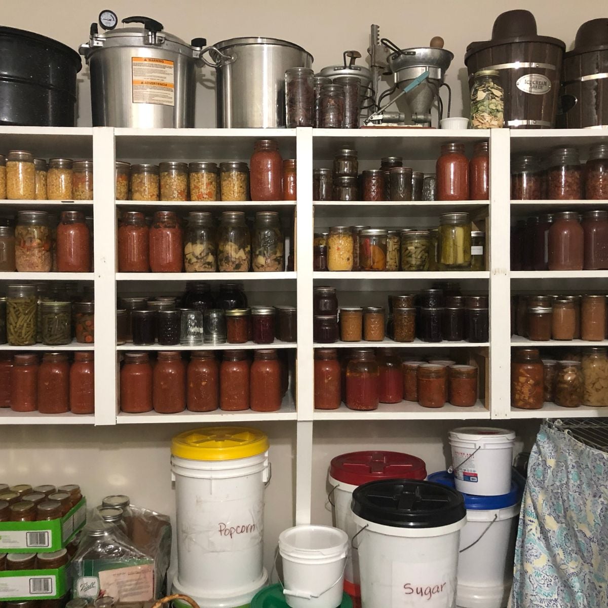 Shelves filled with jars of preserved food, such as pickles and sauces, are organized above large labeled buckets of popcorn and sugar in a pantry. Various kitchen supplies and appliances are on the top shelves.