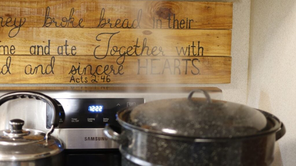 A steaming pot on a stovetop in front of a wooden sign inscribed with “They broke bread in their homes and ate together with glad and sincere hearts, Acts 2:46.”